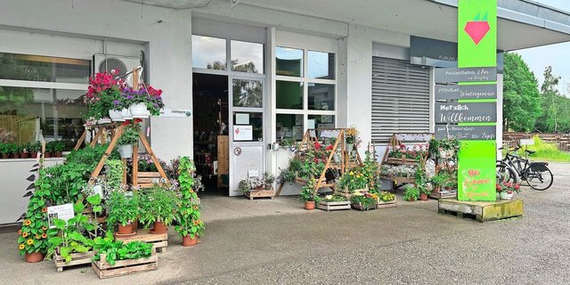Im Eingangsbereich stehen Blumen und Setzlinge zum Verkauf. Trotz des Schildes erinnert der Hofladen an seine frühere Bestimmung als Garage. Von Vorteil ist die grosszügig bemessene Parkplatzzone.