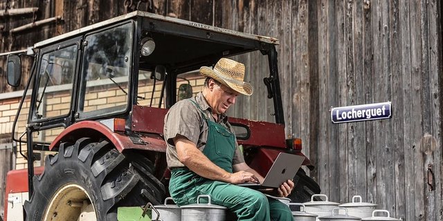 Landwirt sitzt auf Milchkannen und schreibt auf einem Notebook.